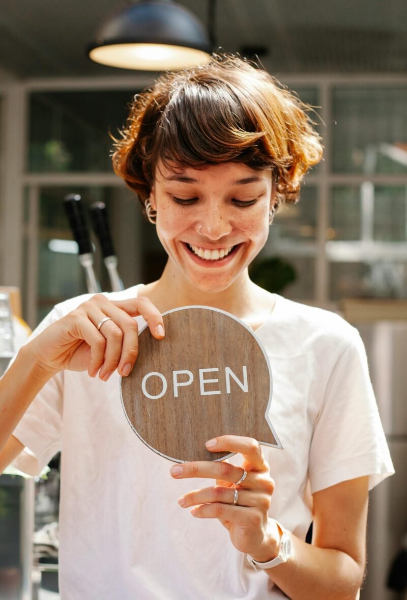 Through glass of happy female worker holding wooden Open sign working in modern cafe with glowing lamp inside